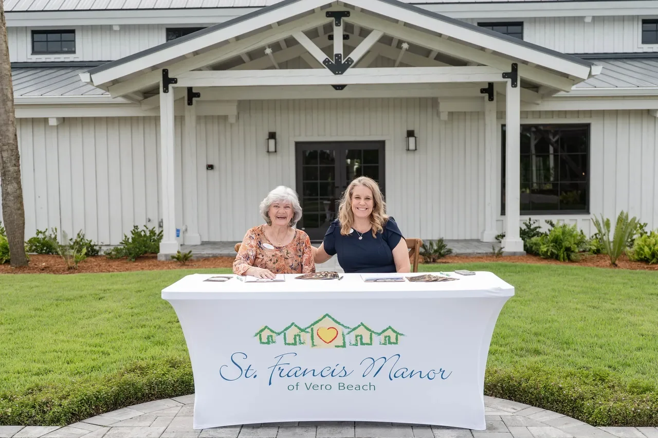 Two women sitting at a table with a St. Francis Manor sign outside a white building with a porch, greenery, and a lawn.