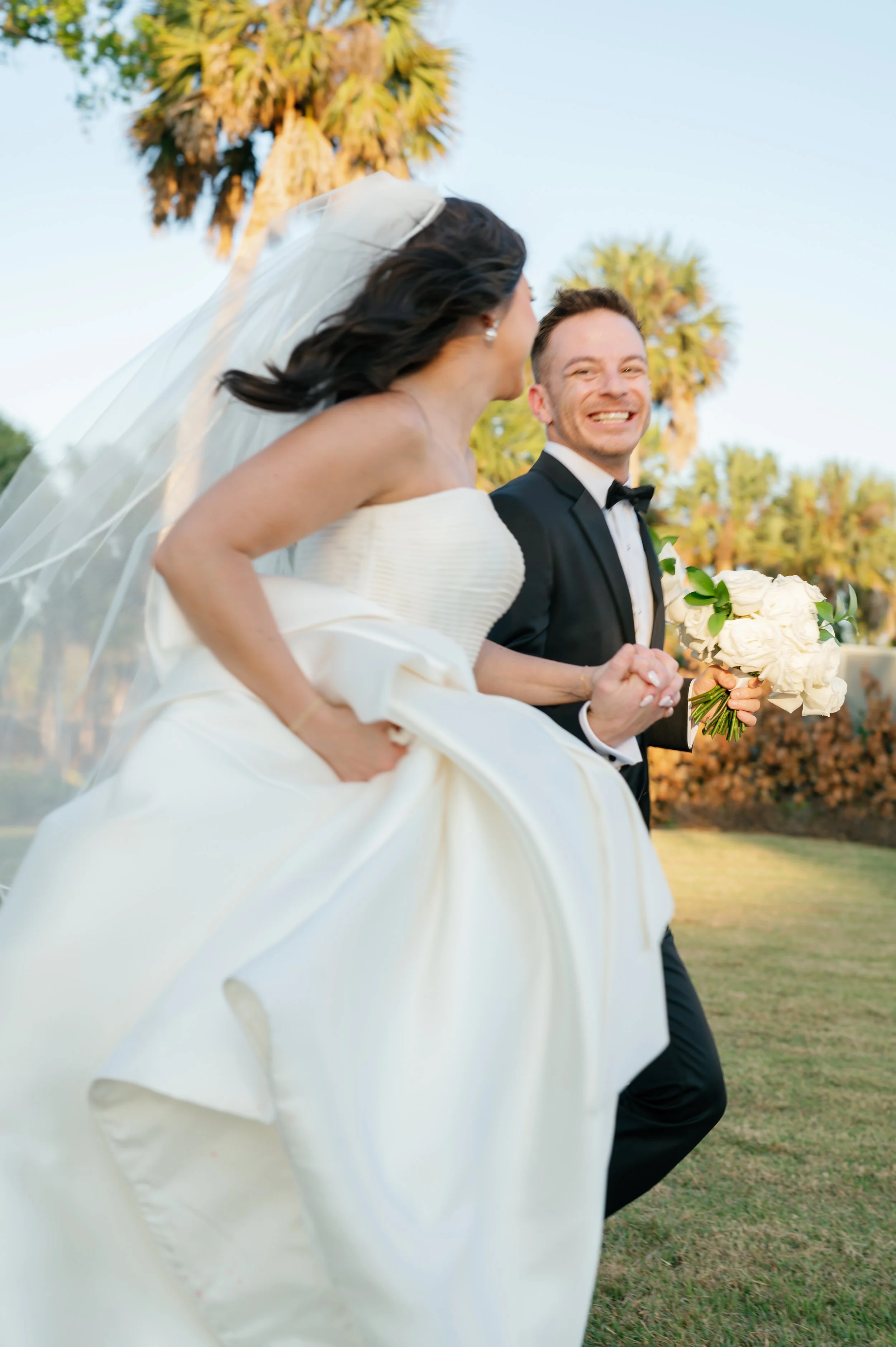 A bride and groom running outdoors during their wedding, with the bride in a white wedding dress and veil, and the groom in a black tuxedo, holding a bouquet of white roses, with palm trees in the background.