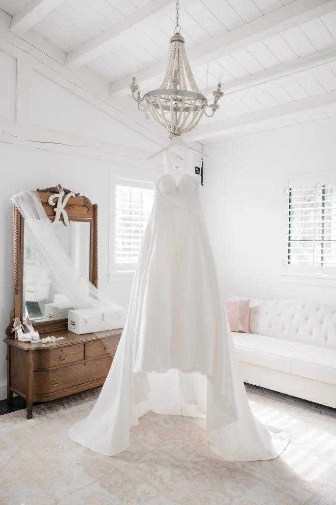 White wedding dress hanging from the ceiling chandelier in a bright, white room with windows, a mirror, and a white sofa.