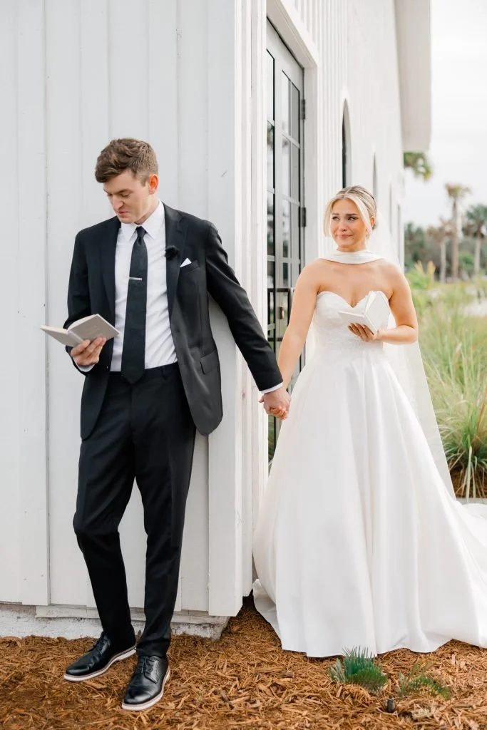 Bride and groom holding hands outside a white building with large windows, the groom is looking at a book while the bride looks into the distance, both dressed in wedding attire.