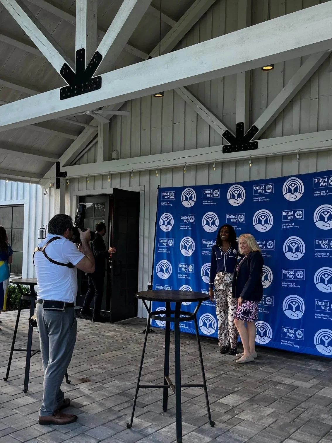 Two women standing in front of a blue United Way backdrop, taking a photo or posing at an event inside a covered outdoor structure with wooden beams and a gray wall. Several people are taking pictures, and there are high tables without chairs in the 