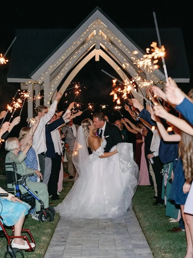 Bride and groom kissing under a decorated archway at night, surrounded by guests holding sparklers during a wedding celebration.