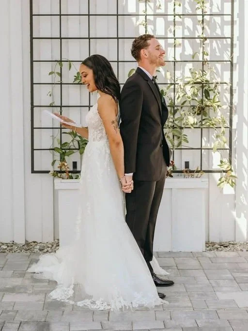 A bride and groom stand back-to-back outside, holding hands. The bride is reading wedding vows, wearing a white lace wedding dress, and the groom is in a black suit and tie. They are smiling with their eyes closed against a white wall with a lattice 
