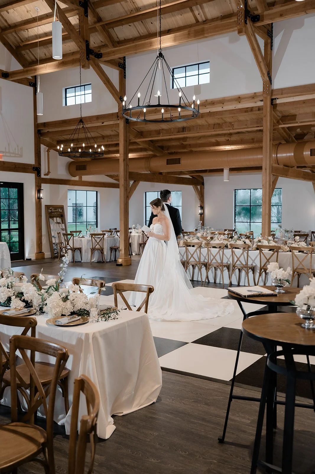 Bride and groom standing during wedding ceremony in a decorated rustic event hall with wooden beams, chandeliers, and floral centerpieces.