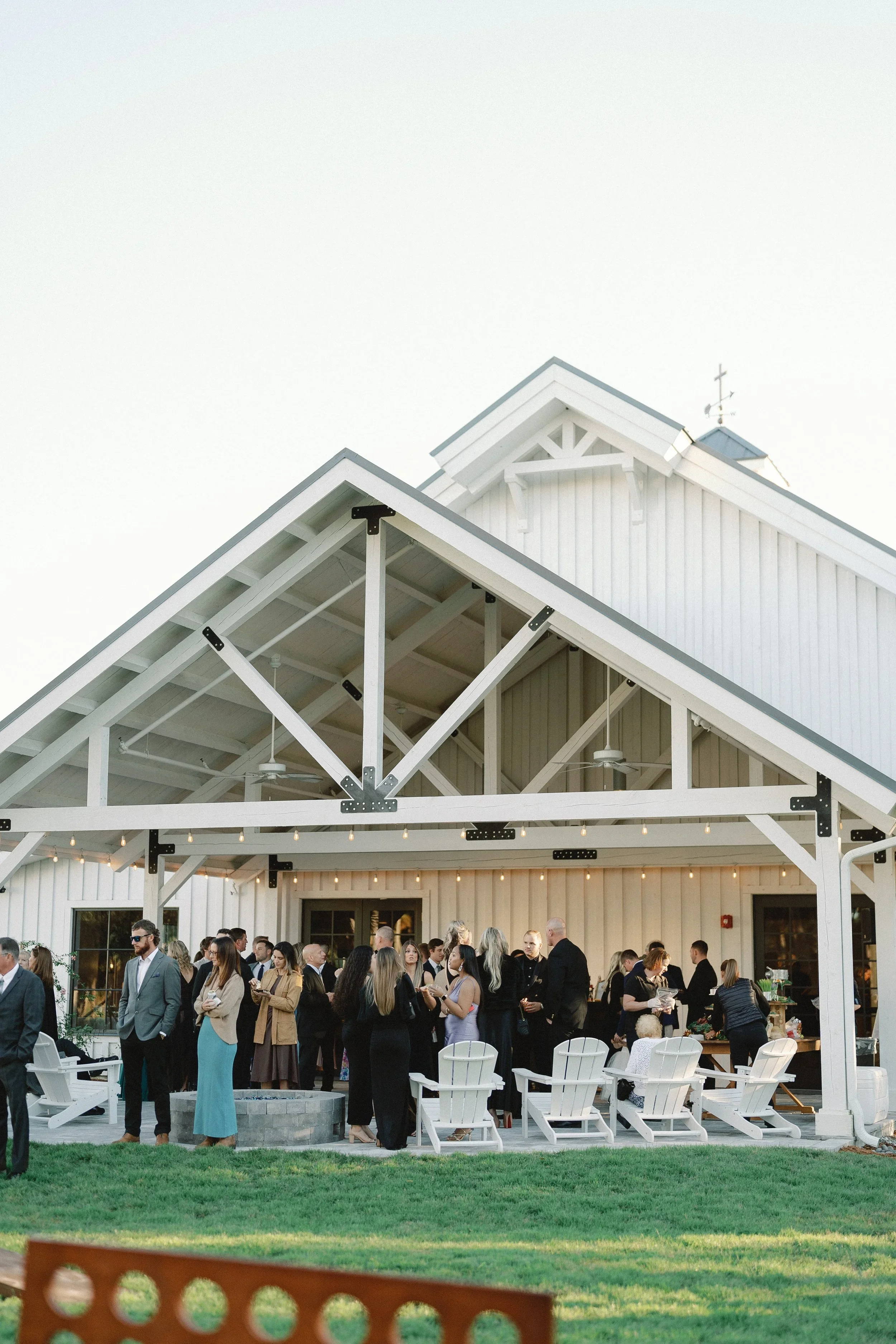 People gathered at an outdoor event under a white barn with string lights, green grass in foreground, and a clear sky.