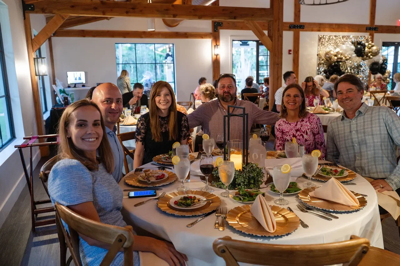 People sitting around a decorated banquet table at a festive gathering in a rustic venue with wood beams and holiday decor.
