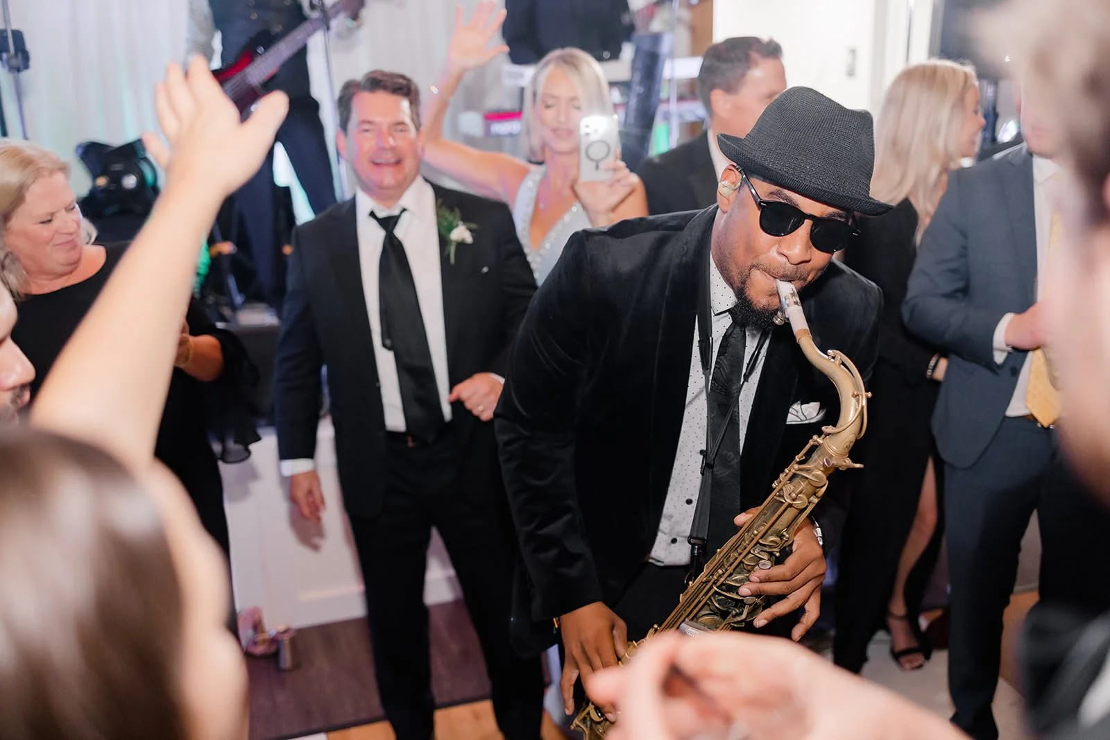 A man wearing a black hat, dark sunglasses, and a black velvet jacket playing the saxophone at a lively party surrounded by people dressed in formal attire, dancing and taking photos.