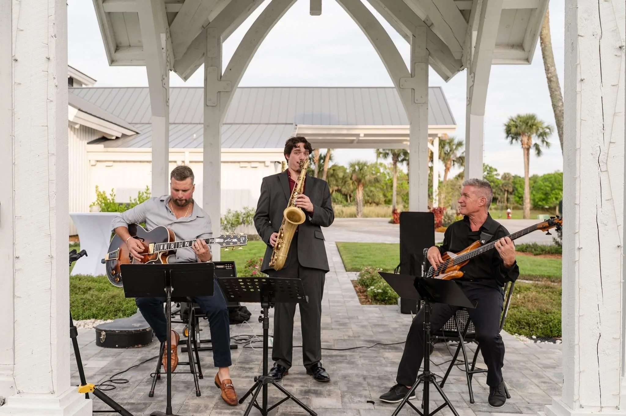 Three musicians performing outdoors under a white wooden pavilion. One playing a guitar, one singing with a saxophone, and one playing a bass guitar. The background shows palm trees and a cloudy sky.
