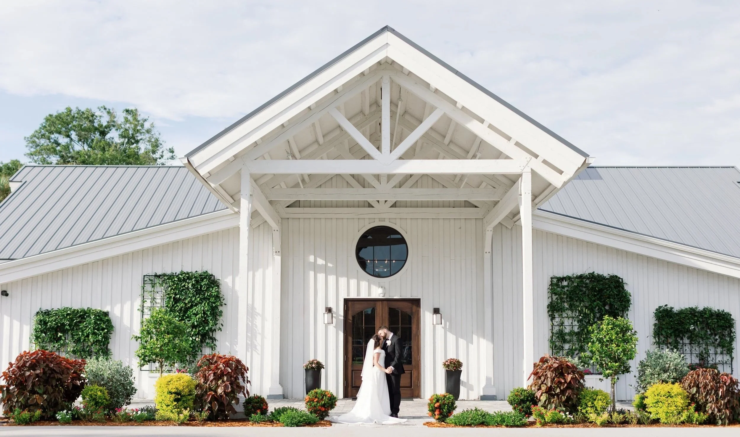 A bride and groom sharing a kiss in front of a white barn at sunset during a wedding ceremony.