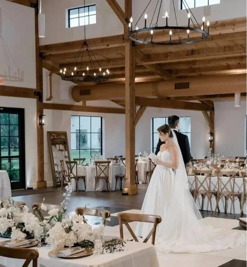 Bride and groom in a rustic wedding reception venue with exposed wood beams, chandeliers, and decorated banquet tables