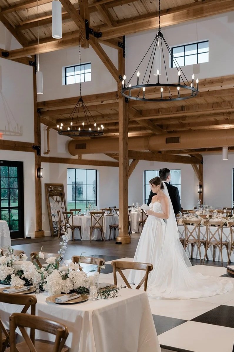 A bride and groom standing in a decorated wedding reception hall with wooden beams, round chandeliers, and tables with white tablecloths and floral centerpieces.