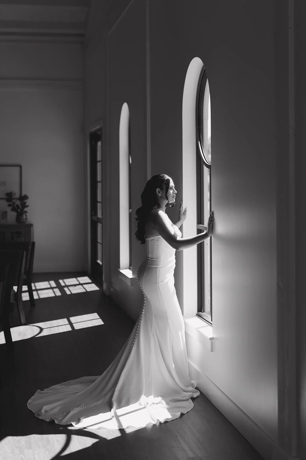 Black and white photo of a woman in a wedding dress looking out a window, with sunlight casting shadows on the floor.