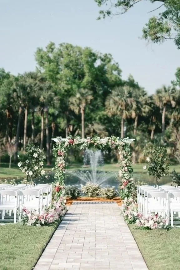Outdoor wedding setup with a brick aisle, white chairs on both sides decorated with pink and white flowers, a floral arch with greenery and pink roses, and a fountain in the background surrounded by trees.