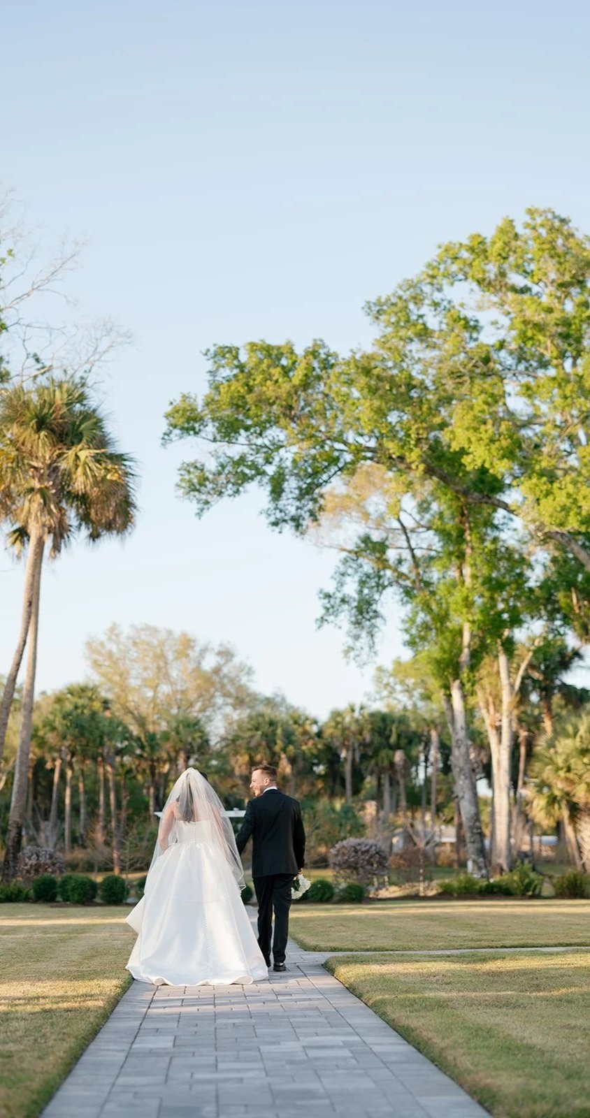 A bride and groom walking hand-in-hand down a garden path, lined with green grass and trees, under a clear blue sky.