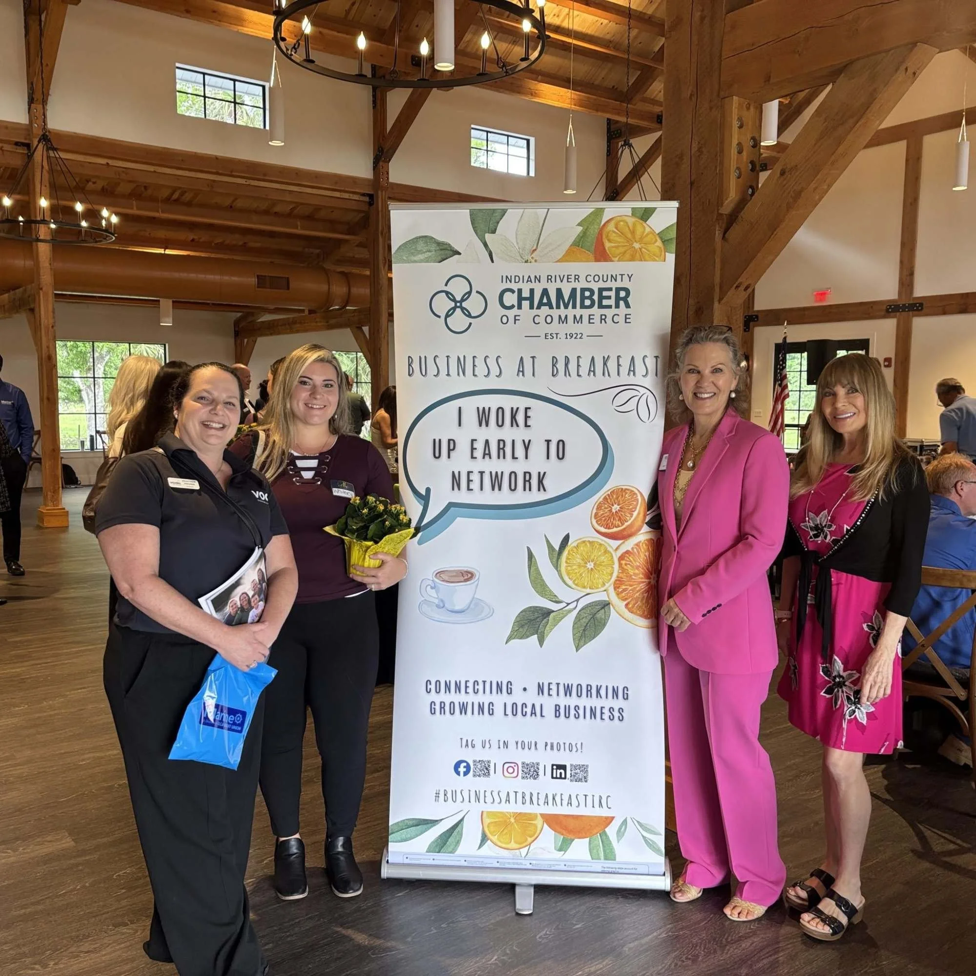 Group of four women standing together at a business breakfast event, smiling, with a tall vertical banner behind them that reads 'Indian River County Chamber of Commerce' and includes graphics of citrus fruits, coffee, and social media icons. The set
