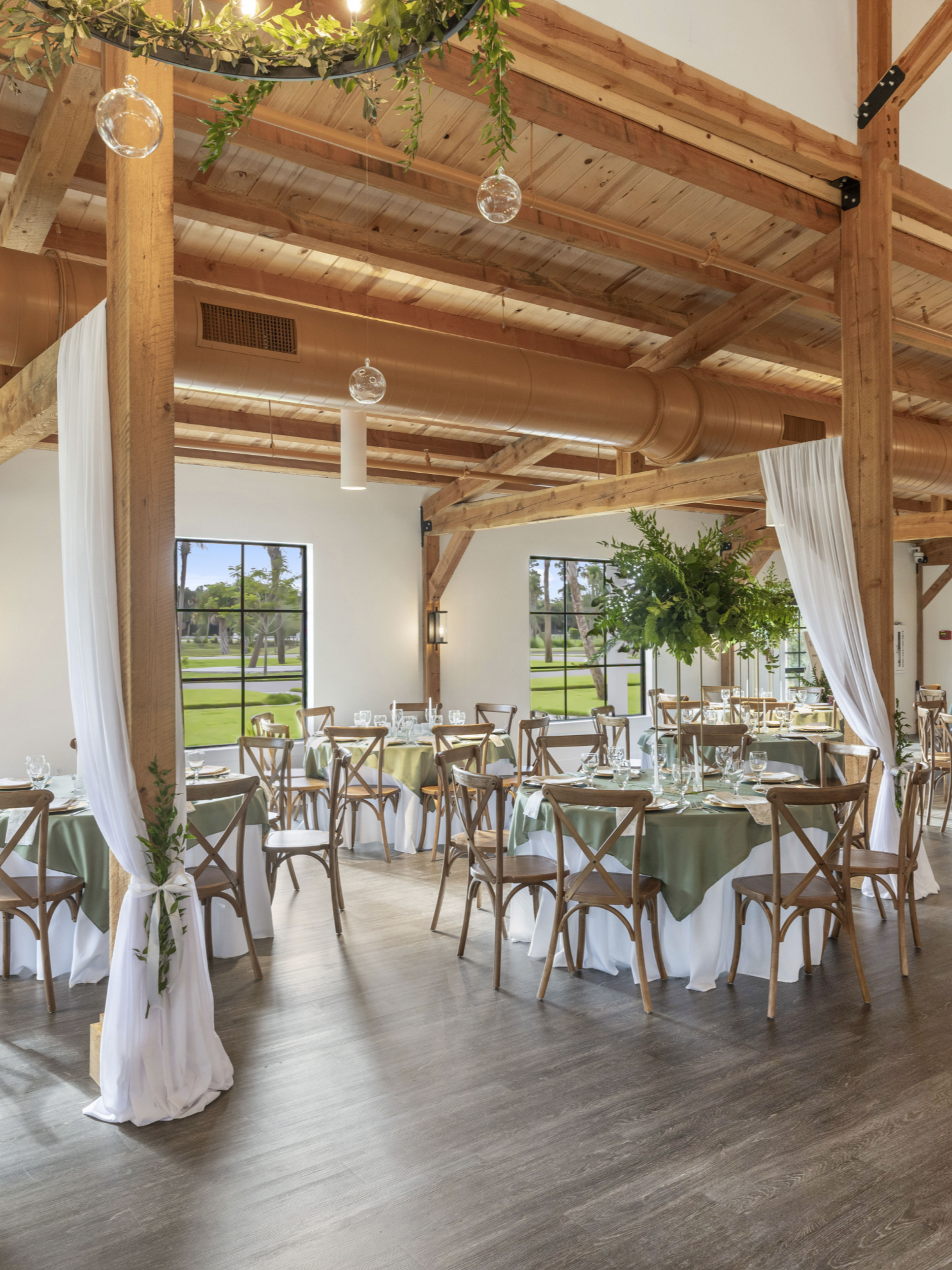 A spacious room decorated for an event with round tables covered in white tablecloths and green overlays, surrounded by wooden chairs. There are white drapes tied to posts with greenery accents. Large windows let in natural light, showing a view of trees and grass outside. The ceiling has exposed wooden beams, ductwork, and hanging glass light fixtures.