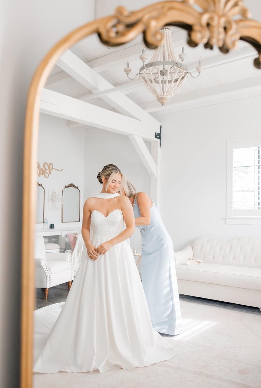 A bride in a white wedding gown standing in a bright, elegant room with white walls and furniture. She is smiling with closed eyes, while a woman in a light blue dress adjusts her dress.