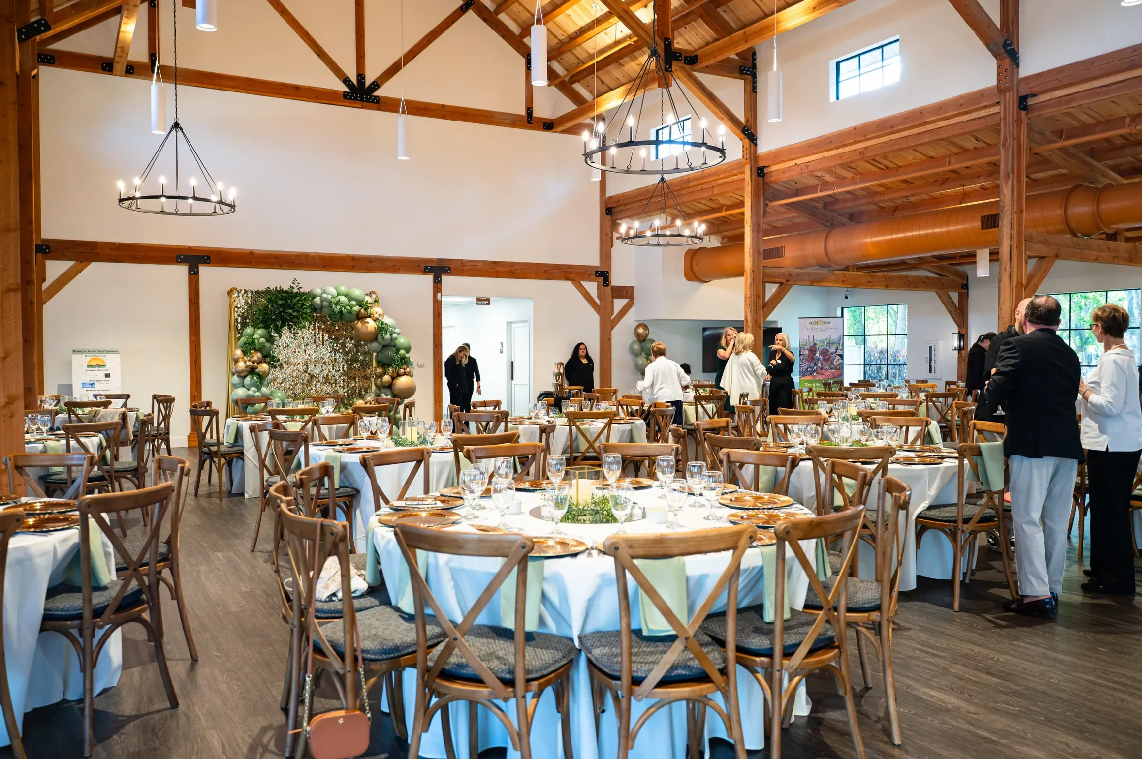 Interior of a large event hall with high wooden ceilings, round tables with white tablecloths, and place settings including glassware and gold-colored plates. There are several guests and staff members standing and chatting, with a balloon decoration