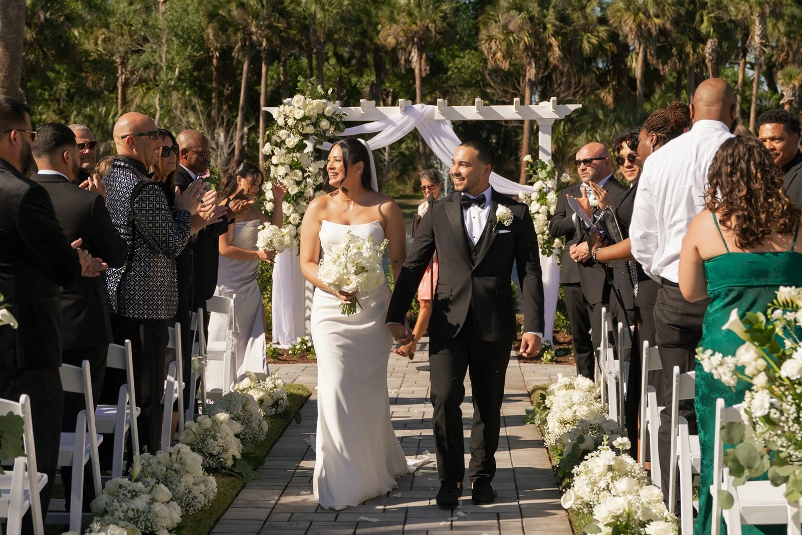 A bride and groom walk down the aisle holding hands at an outdoor wedding ceremony. The bride wears a white strapless gown, and the groom is in a black tuxedo. Guests on both sides applaud them.