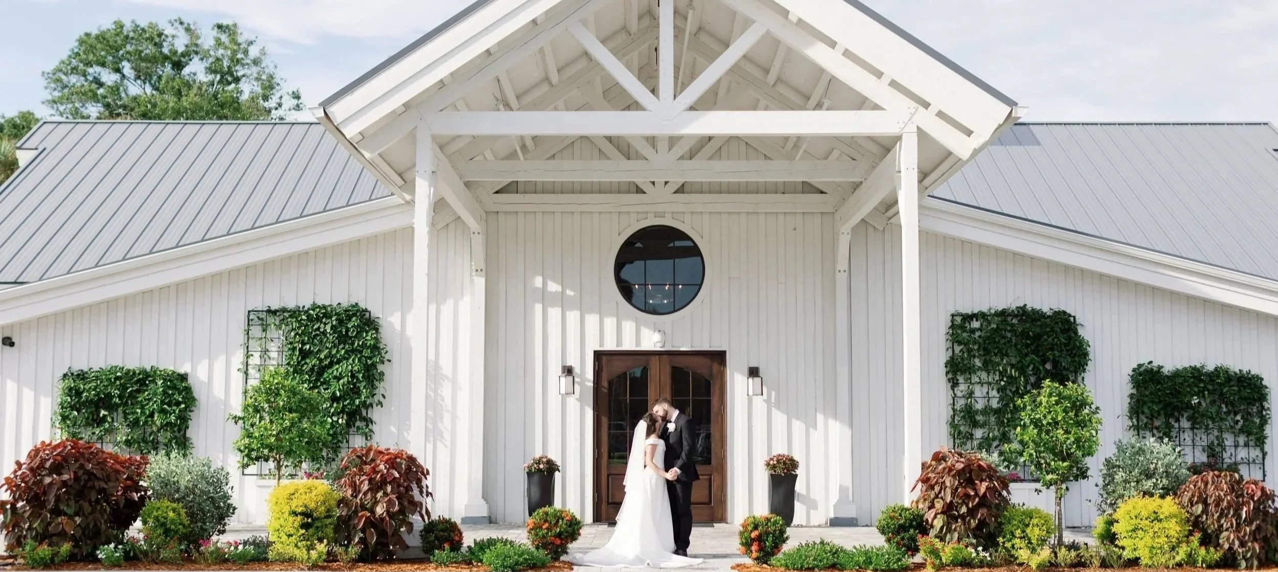 A bride and groom sharing a kiss outside a white barn decorated with greenery and flowers.