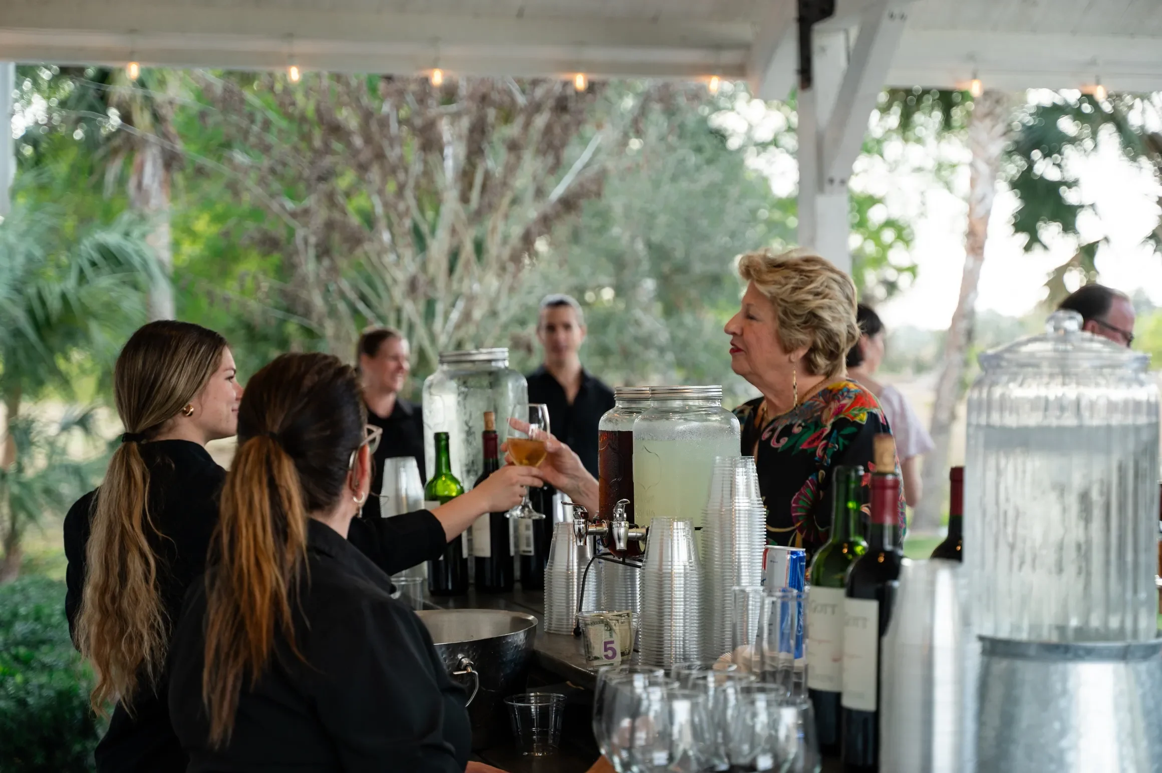 Women working at a bar with various bottles and glasses, surrounded by greenery and string lights.