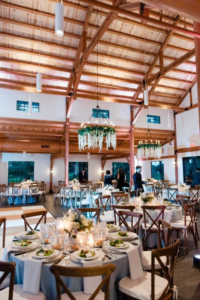 A decorated event hall with round tables set with plates, glasses, and floral centerpieces, and hanging chandeliers with greenery; the hall has a wooden ceiling and large windows.
