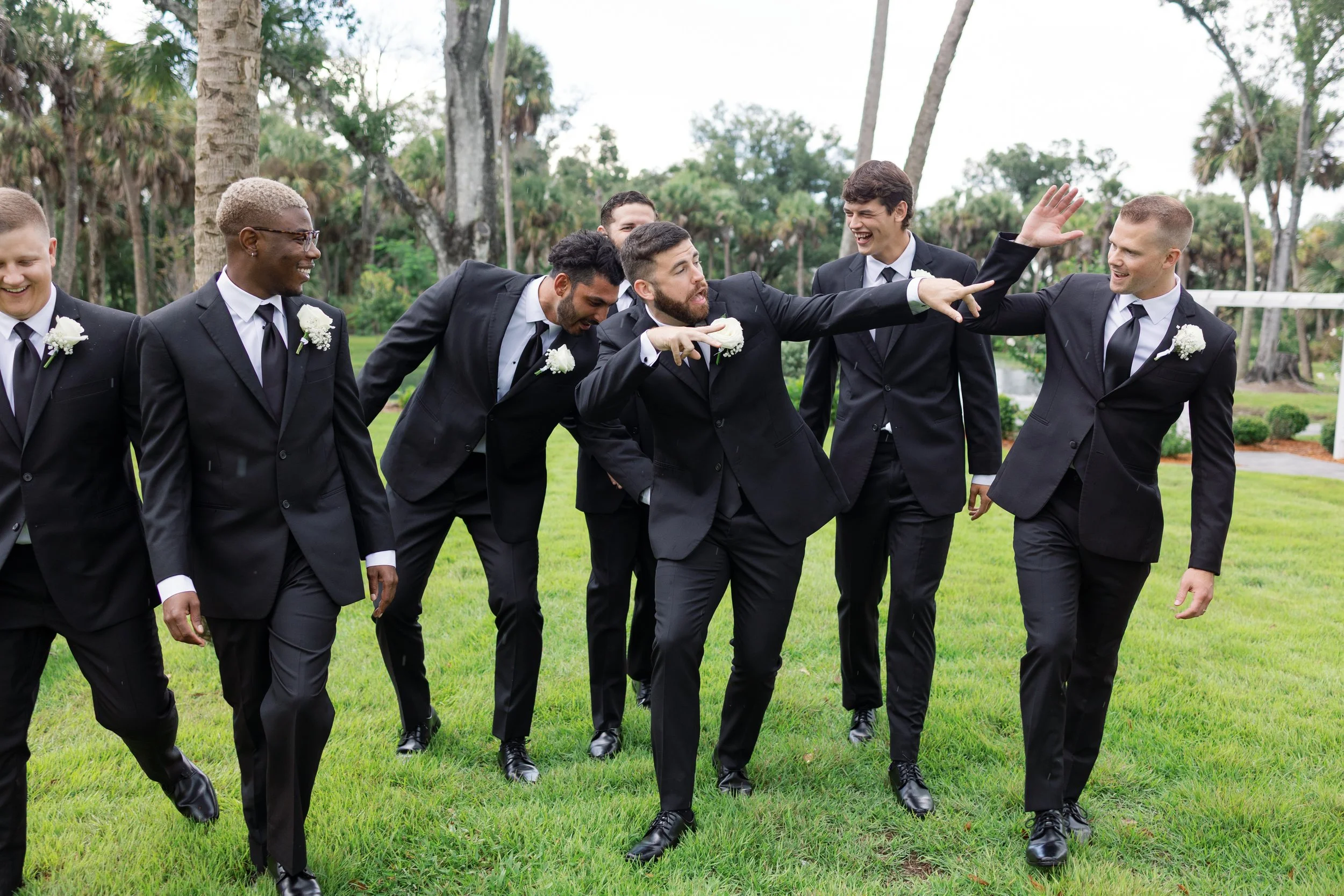 Group of men dressed in black suits and white shirts, laughing and walking on a grassy lawn with trees in the background.