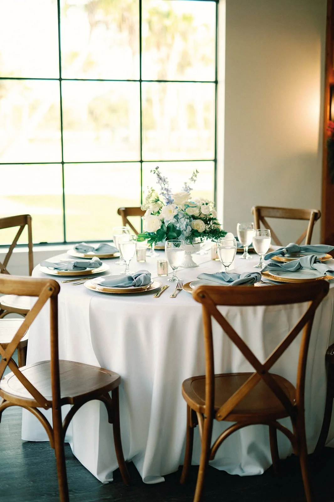 Round banquet table set for six, with a white tablecloth, gold-rimmed plates, blue napkins, silverware, wine glasses, and a white floral centerpiece, in a room with large window and natural light.