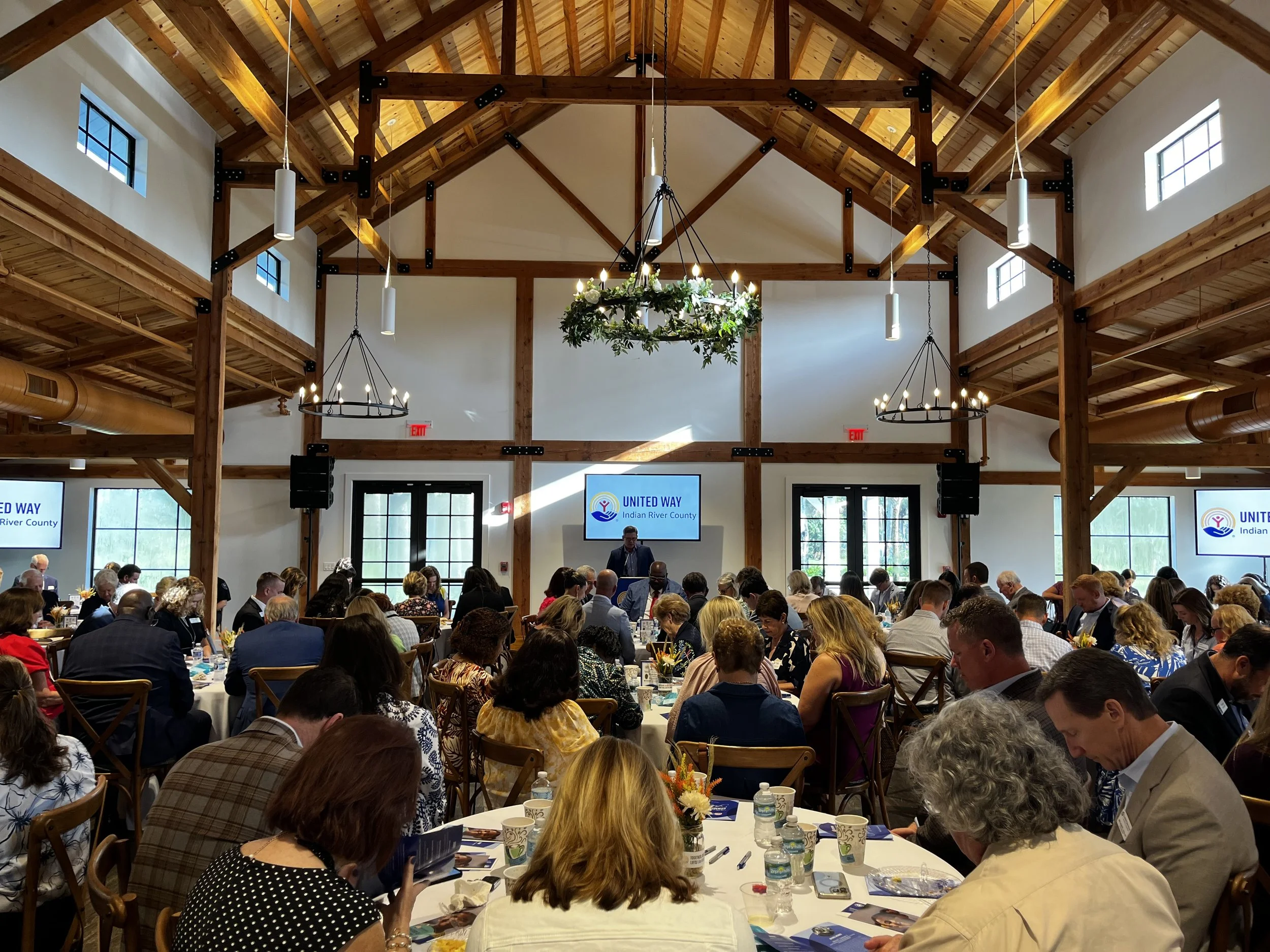 A large indoor event with many people seated at round tables in a spacious, wooden ceiling hall. There are large windows, chandeliers with candles, and a wreath-style light fixture hanging from the ceiling. The event appears formal or community-orien