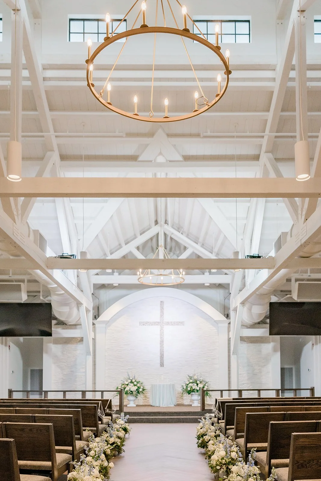 Interior of a church decorated for a wedding with white flowers along the aisle, a cross on the white wall at the altar, and chandeliers hanging from the ceiling.