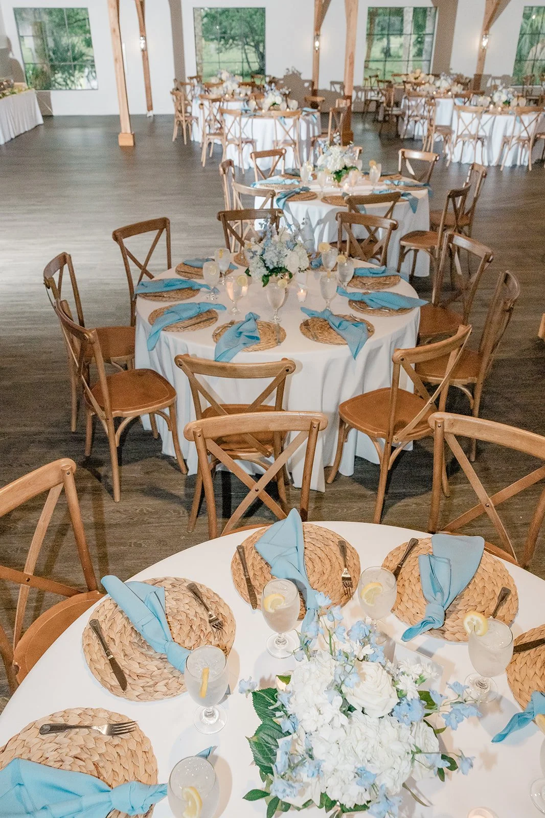 Elegant wedding reception setup with round tables draped in white tablecloths, decorated with light blue napkins, glassware, and floral centerpieces, surrounded by wooden cross-back chairs in a spacious, well-lit venue with large windows and wooden beams.