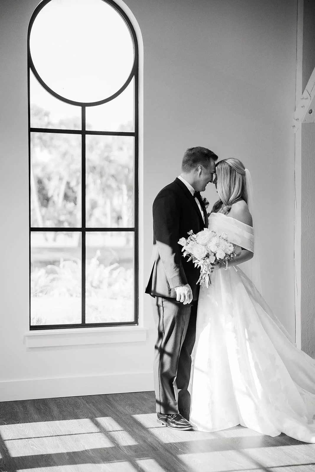 A black and white photo of a bride and groom standing close with foreheads touching, holding a bouquet, near a window with sunlight casting shadows on the wooden floor.