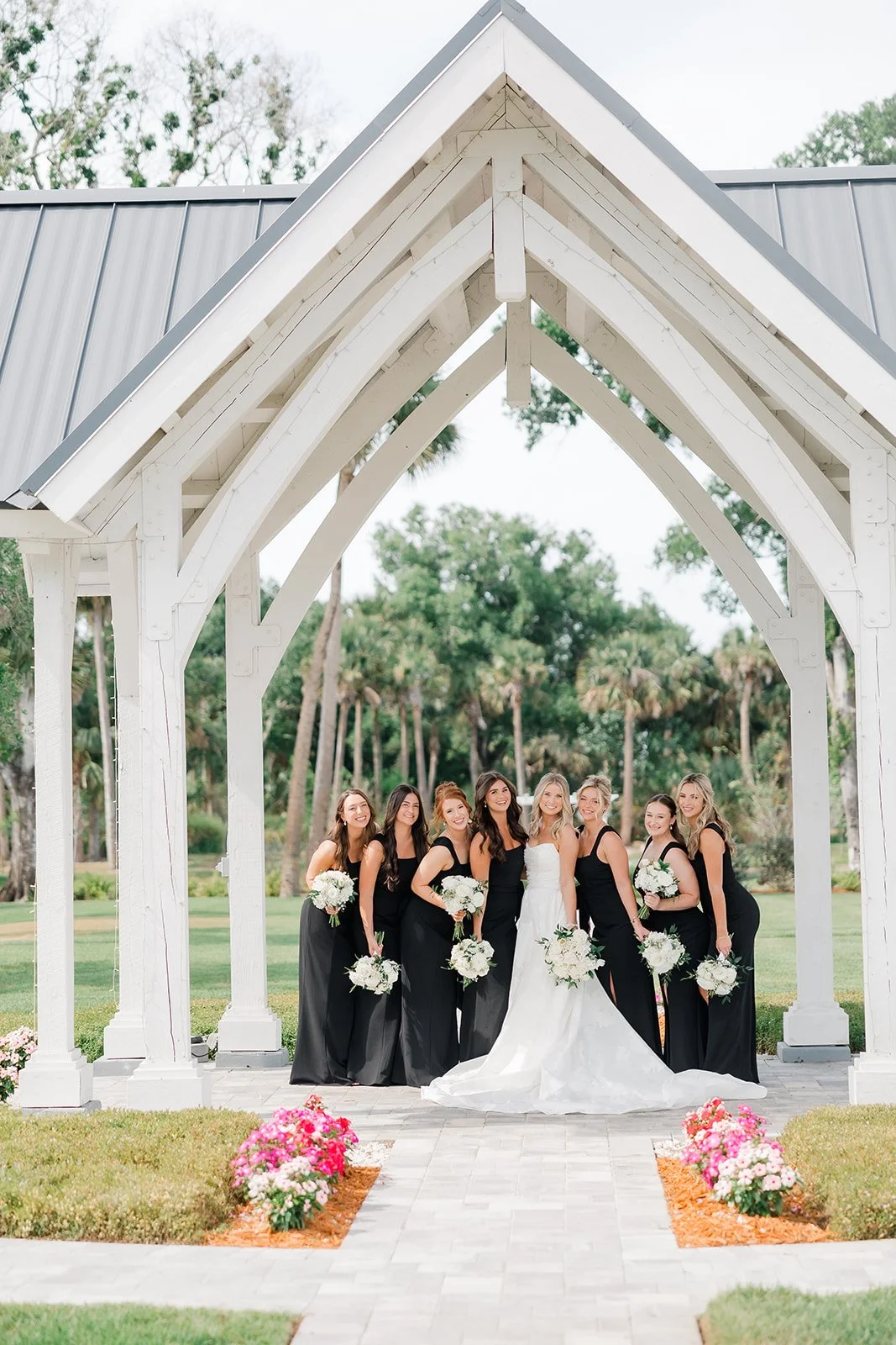A group of women in formal dress standing under a white wooden arch with greenery and palm trees in the background, holding bouquets.