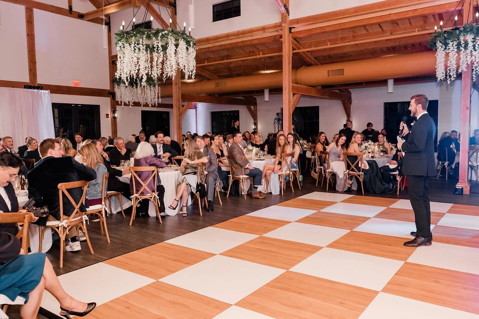 A man in a suit giving a speech at a wedding reception with rows of seated guests watching him, decorated with floral chandeliers and wooden beams.