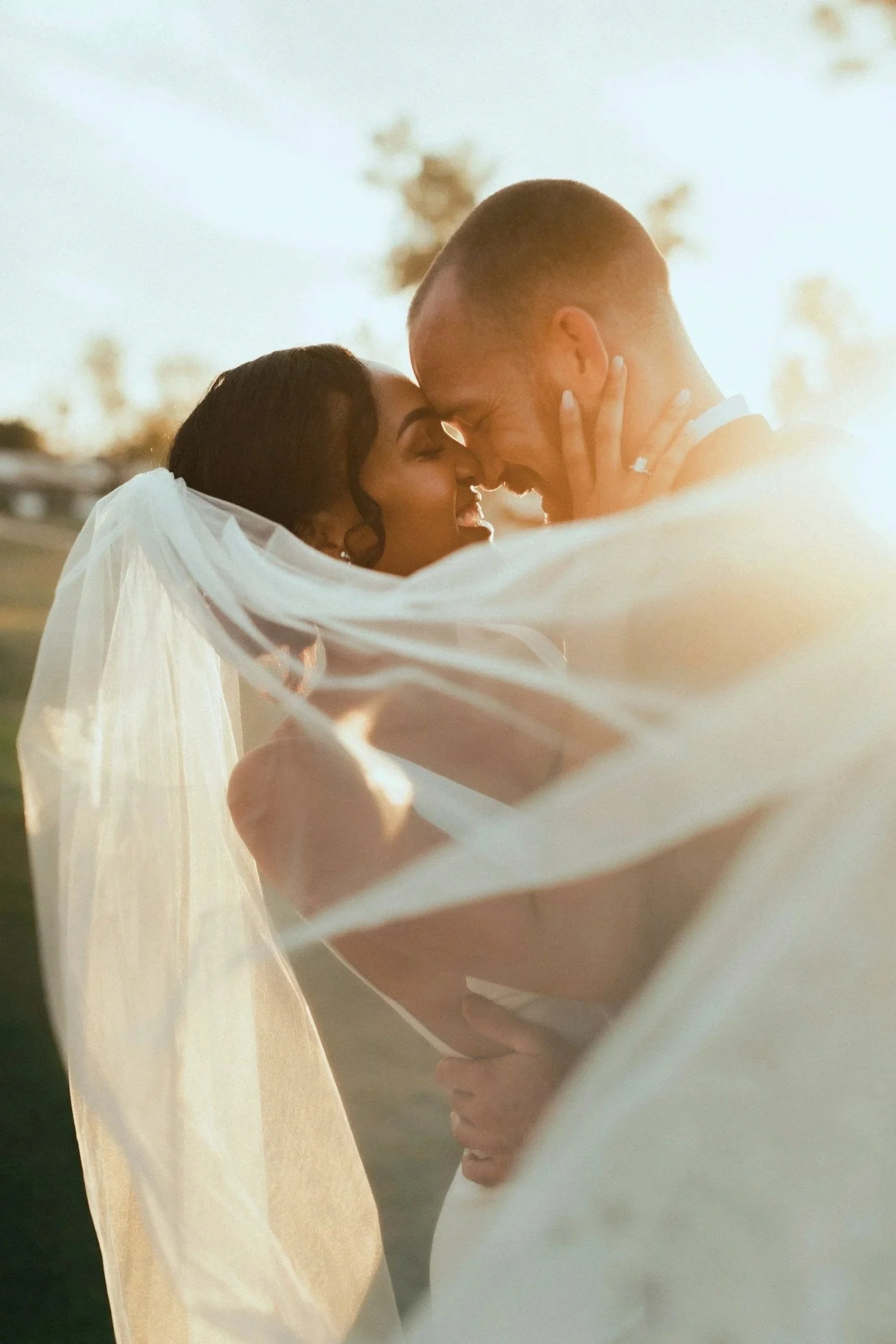 A bride and groom embrace and touch foreheads during their wedding, with soft sunlight illuminating them and festive round lights in the background.