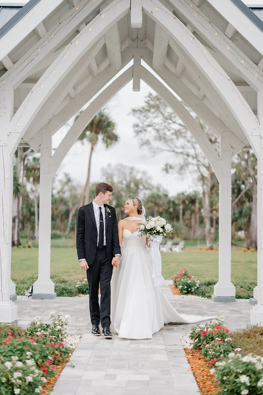 A bride and groom are walking hand in hand under a white wooden wedding arch, with flowers along the pathway, outdoors on a cloudy day.