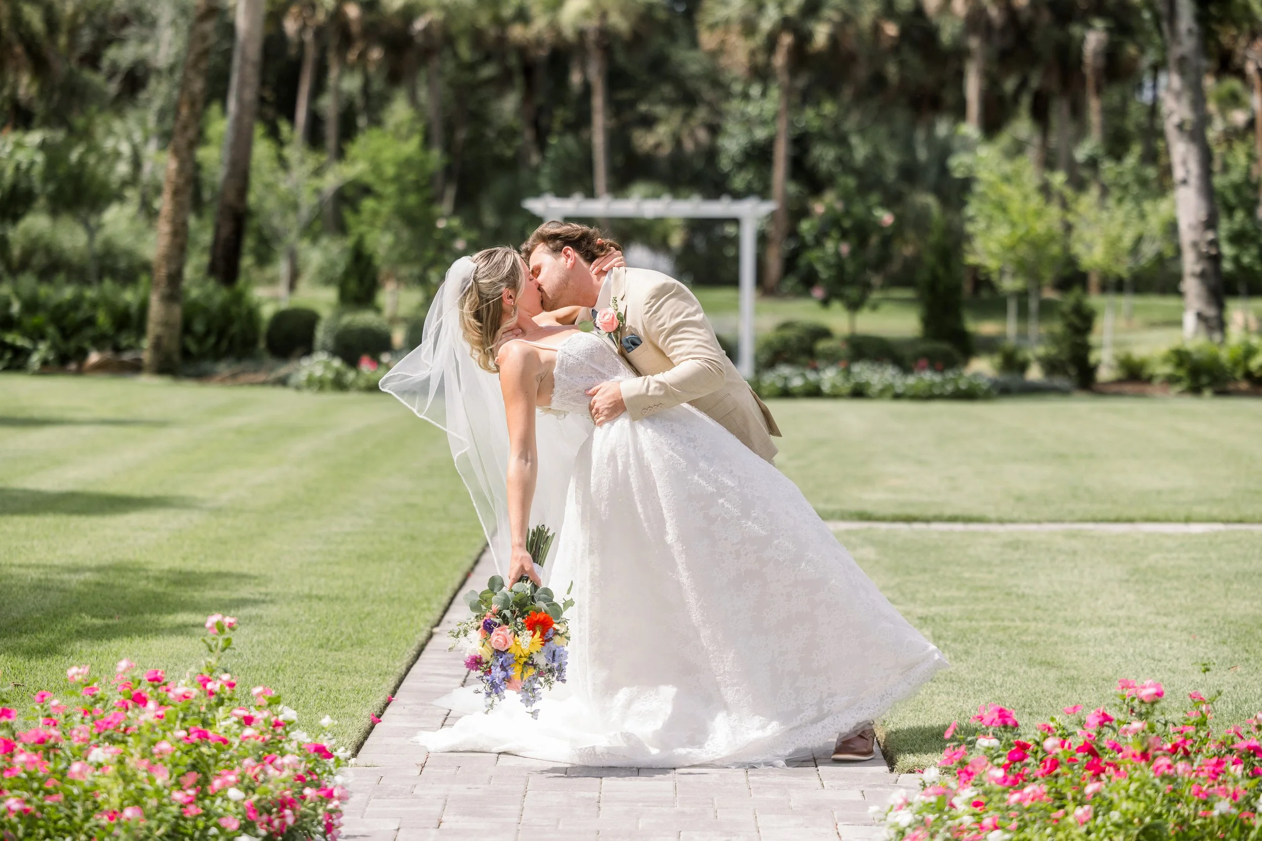 A bride and groom sharing a kiss outdoors during their wedding, with colorful flowers lining the walkway and trees in the background.