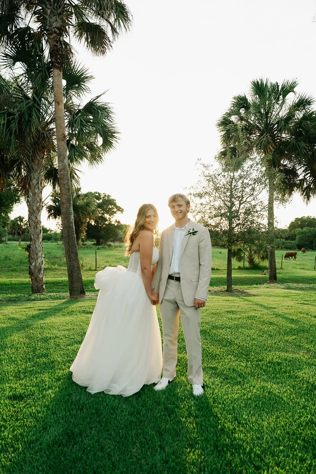 A bride and groom stand hand-in-hand on a lush green lawn with palm trees in the background, during sunset.