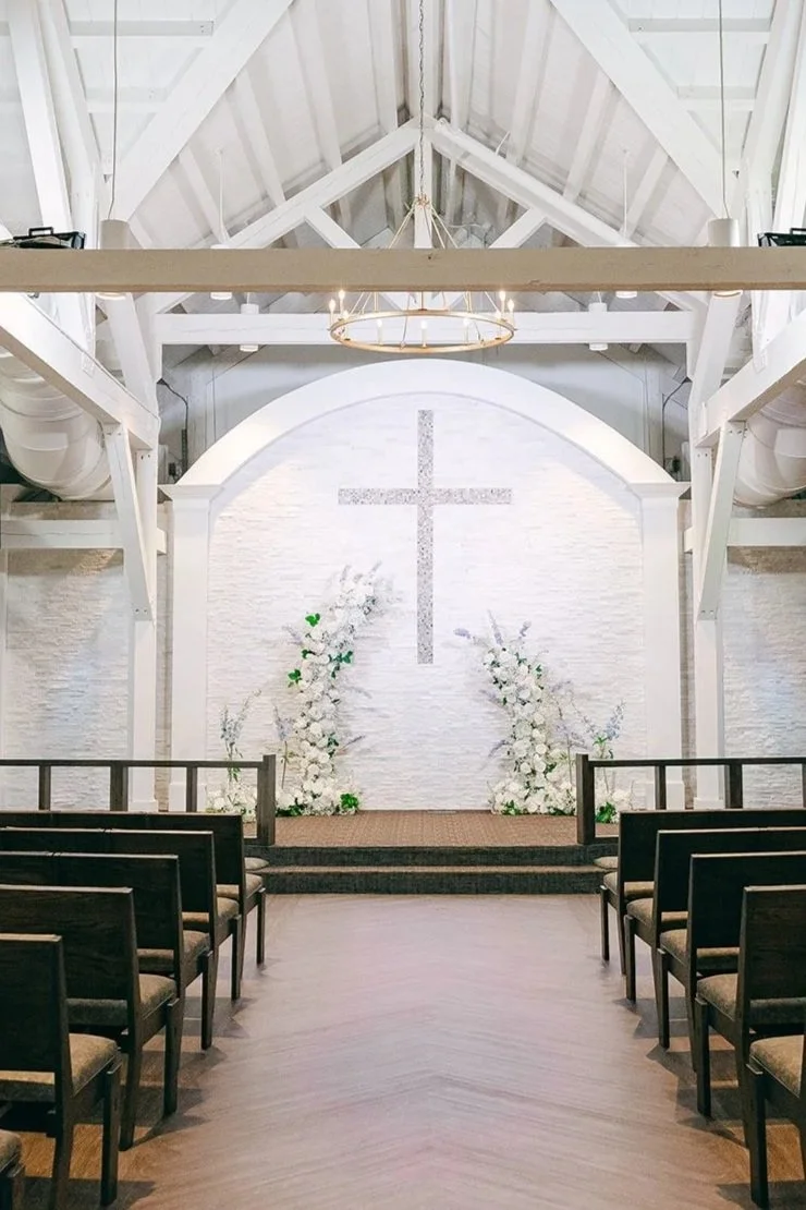 Empty wedding chapel with a white cross on the back wall, floral arrangements on either side of the stage, and rows of chairs facing the altar.