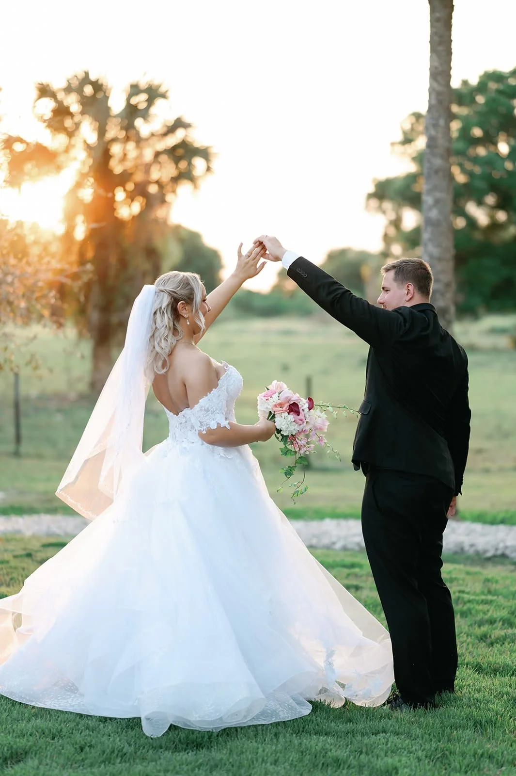 A bride and groom dance outdoors during sunset, with the bride in a white wedding gown holding a bouquet, and the groom in a black suit, raising her hand.
