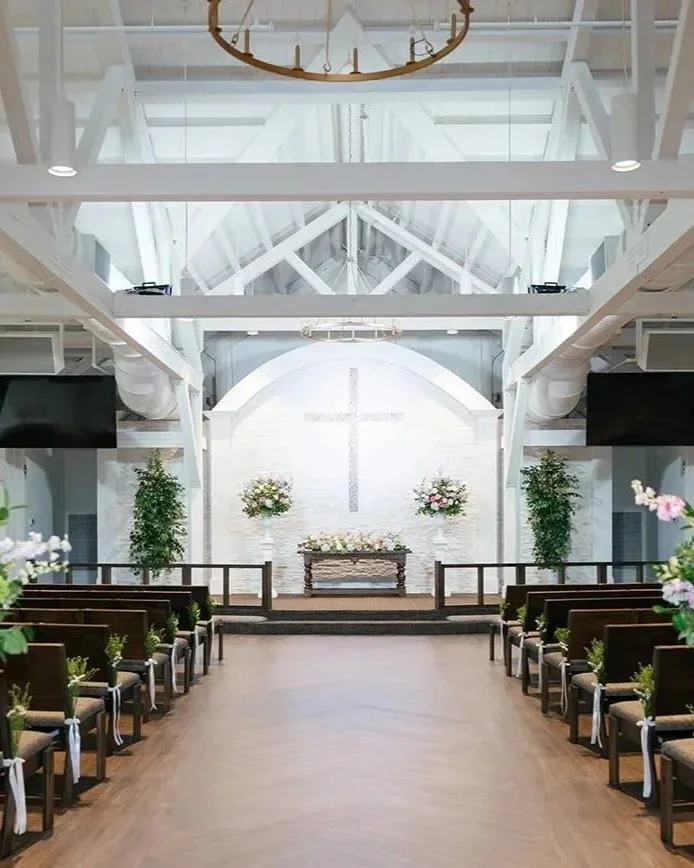 Empty church with wooden pews decorated with flowers, a cross on the white brick wall at the front, and floral arrangements on either side of the altar.