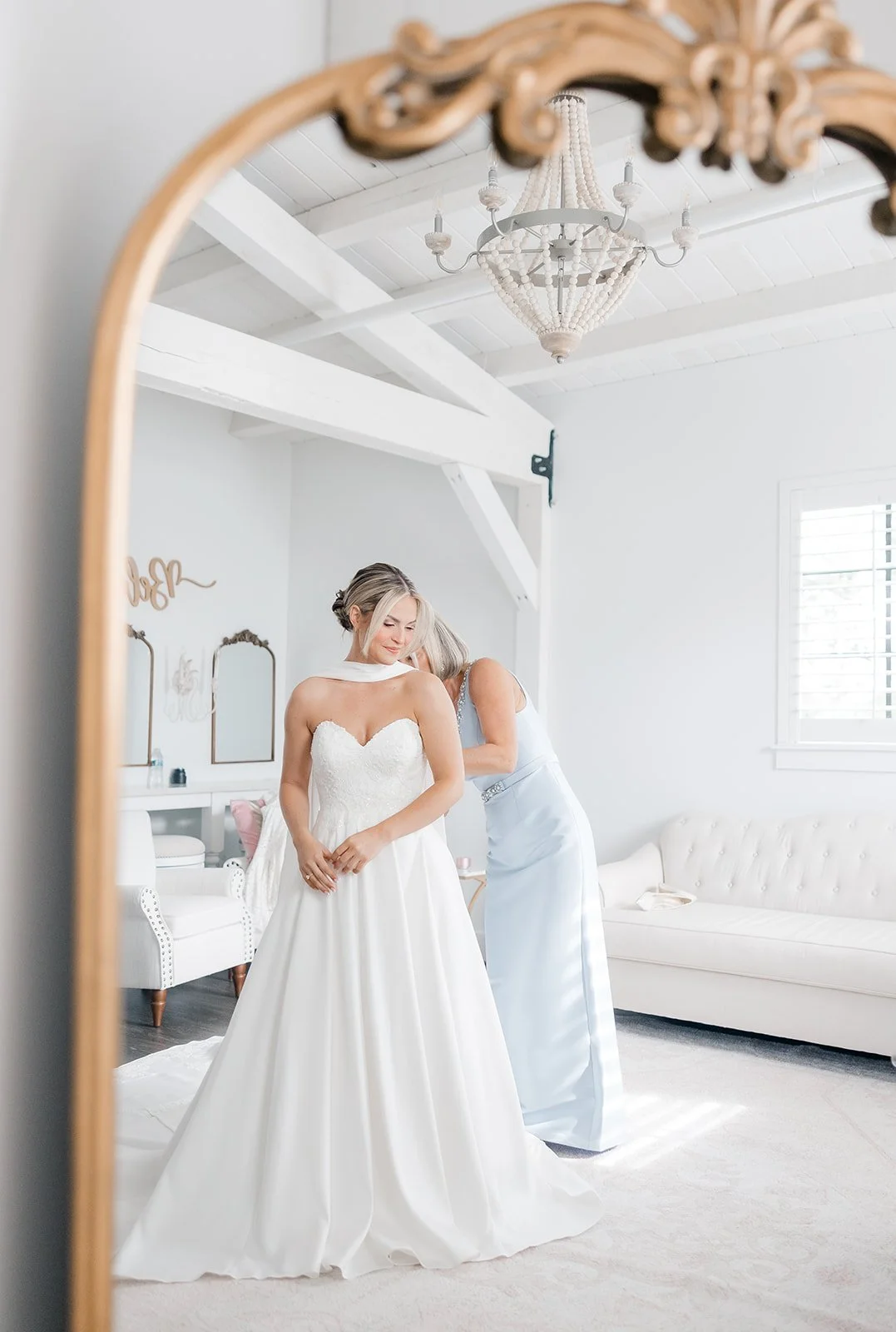 A bride in a white wedding gown being assisted by a woman in a light blue dress inside a bright, white room with a chandelier and a vintage mirror