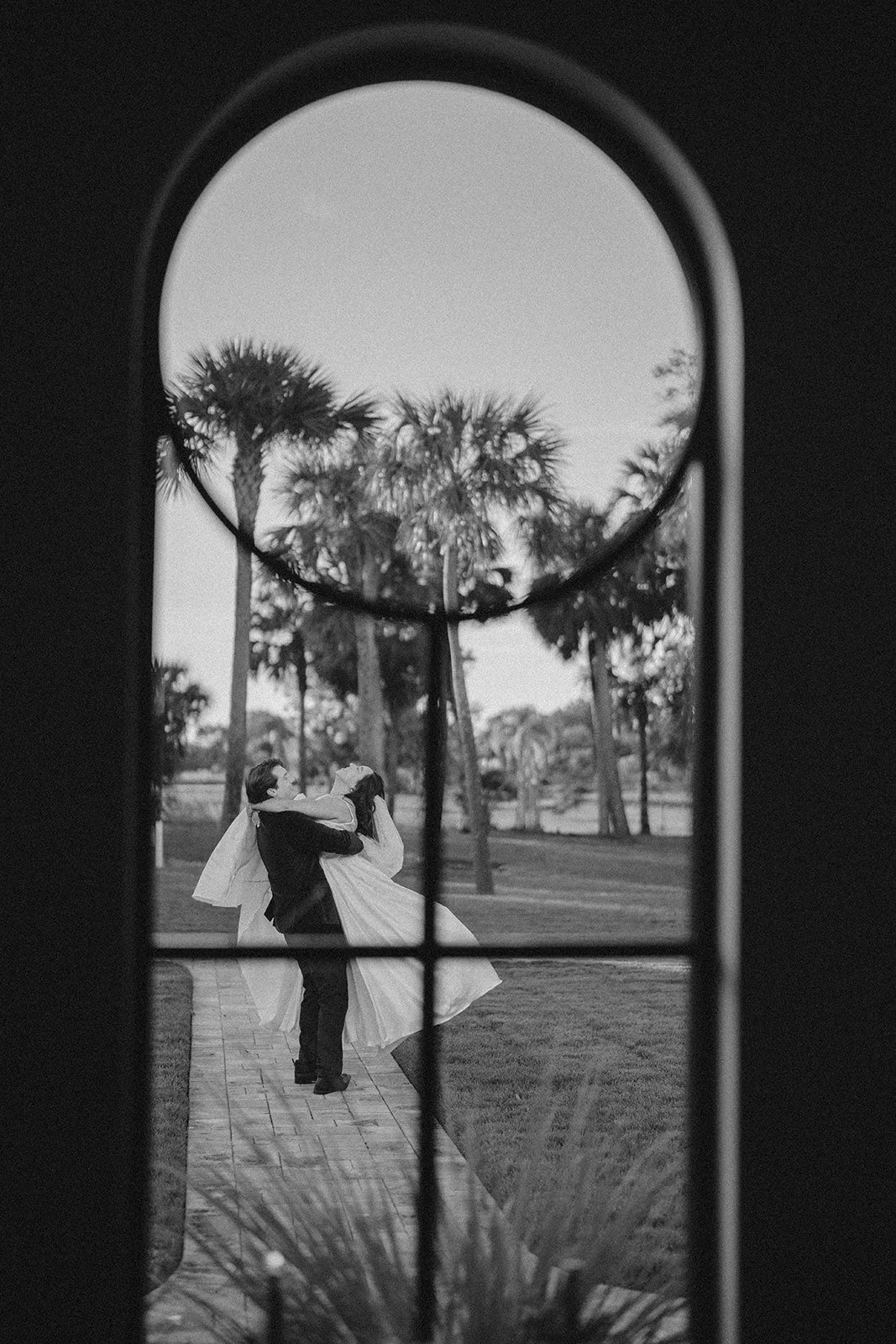 A black and white photo of a couple dancing outdoors viewed through a window with a rounded top. They are surrounded by palm trees and a grassy area.