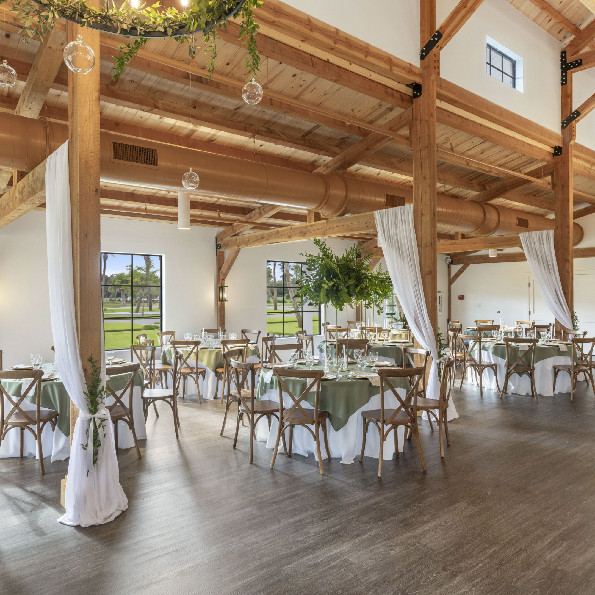 A spacious indoor event space with wooden beams and large windows, set up with round tables covered in light green tablecloths, surrounded by wooden chairs, and decorated with glassware and table settings. White drapes hang from the wooden beams, and there are hanging glass sphere lights and green plants for decoration.