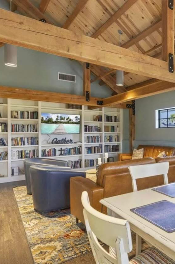 Living room with wooden ceiling beams, a large bookshelf filled with books, a leather sofa, armchair, dining table with placemats, and a rug on the floor.