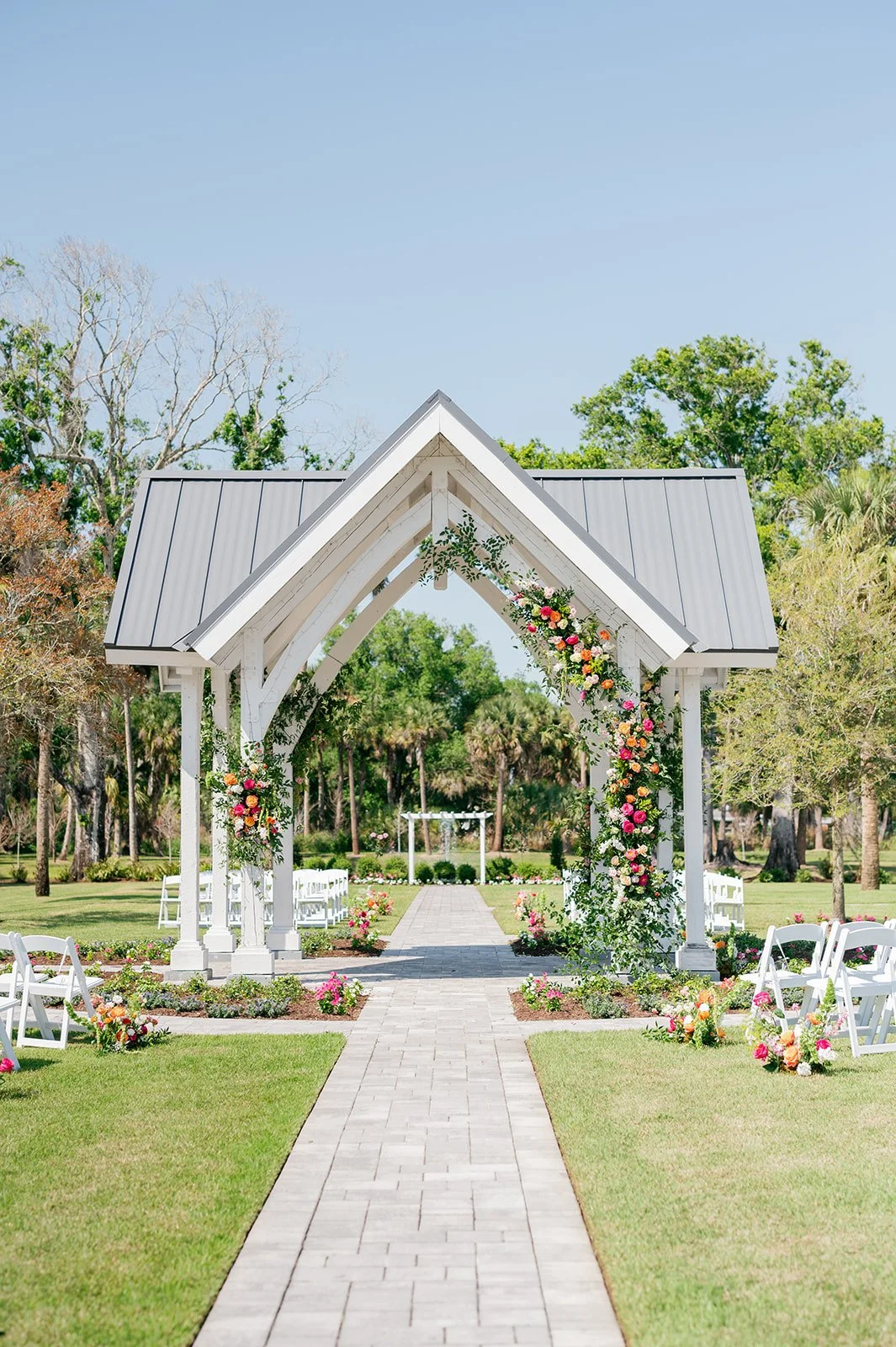 Wedding outdoor ceremony setup with a white wooden arch decorated with flowers and greenery, white chairs along a brick pathway, surrounded by trees and greenery under a clear blue sky.