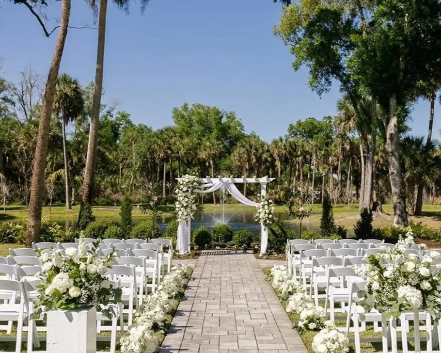 Outdoor wedding setup with white chairs, floral arrangements along the aisle, a decorated arch over a water fountain, and lush green trees in the background on a sunny day.