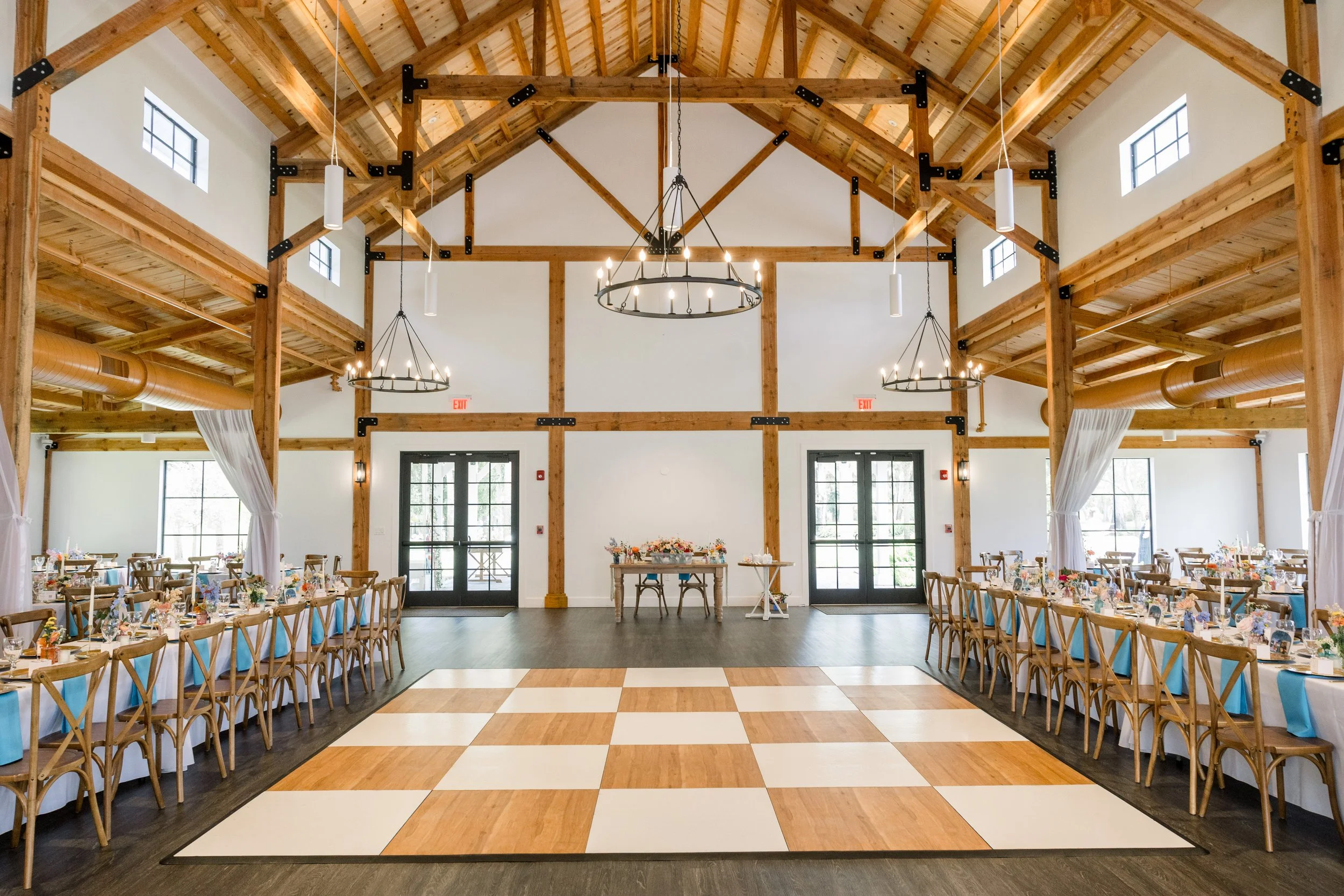 A spacious event hall with a high wooden ceiling, chandeliers, and large windows. The hall is decorated for a celebration with tables set with centerpieces and surrounded by wooden chairs. There are double doors at the back and a checkered dance floor in the center.