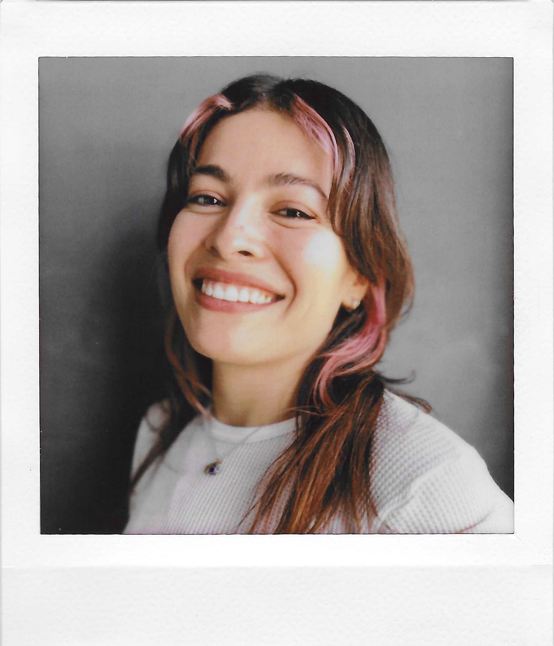 Close-up photograph of a woman smiling, with medium-length hair dyed pink at the tips, wearing a white top, against a gray background.