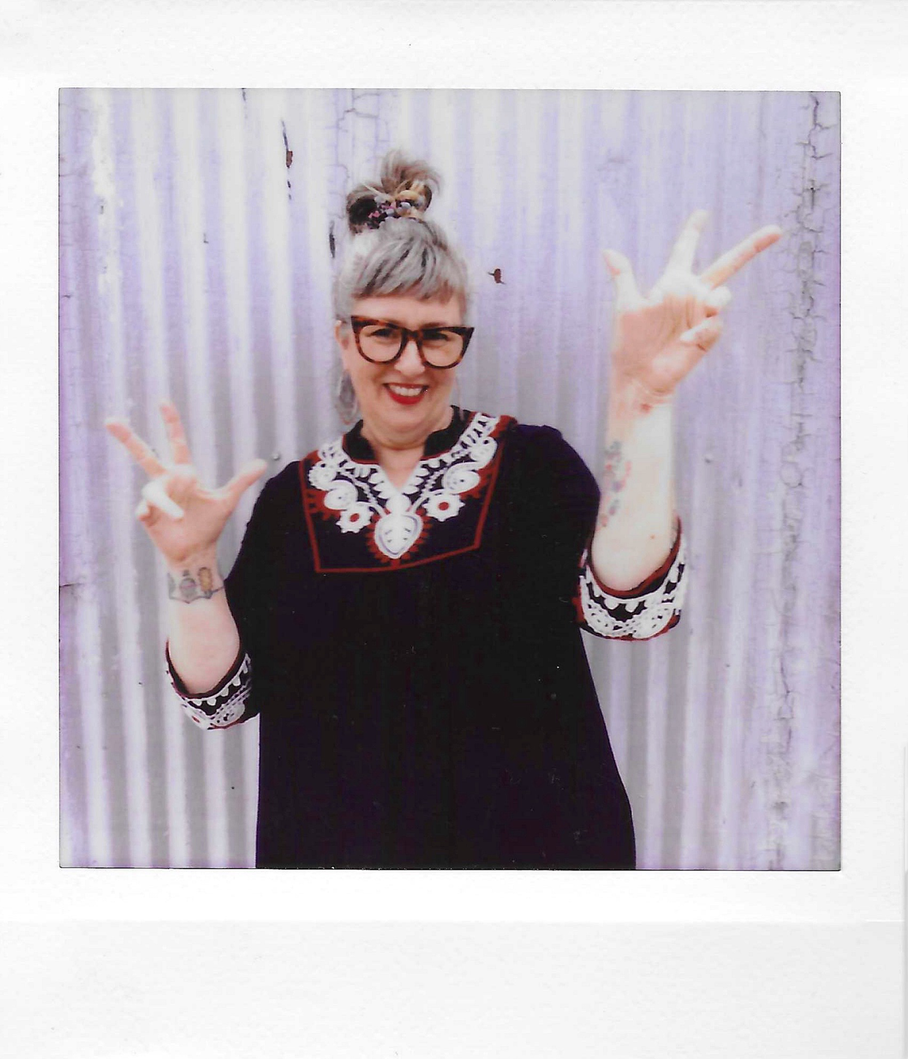 A person with short gray hair, wearing glasses, smiling, and making peace signs with both hands, standing in front of a cracked, weathered wall.